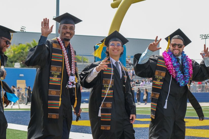 Three College of Science and Health graduates wave at the camera as they walk to their seats at Commencement, 